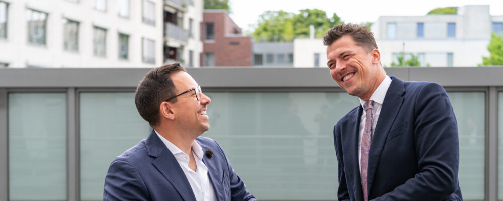 Zwei Männer im Anzug stehen im Freien vor einem modernen Gebäude und lachen miteinander; das Bild begleitet die Stellenanzeige für Lohnbuchhalter (m/w/d) bei der Kanzlei Weber Steuerberatungsgesellschaft mbH in Köln.
